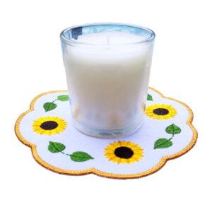 Glass candle displayed on embroidered sunflower candle mat with scalloped edge, yellow blooms, green leaves, and golden-yellow border, adding warmth and rustic charm to home décor.