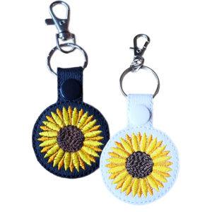 Sunflower keyrings shown side by side, one in black and one in white, each featuring a circular sunflower design and attached to a silver keyring ring with a lobster clasp.