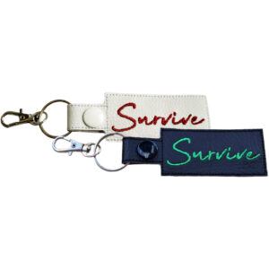 Two rectangular keyrings with the word "Survive" embroidered in cursive. One keyring is black with green embroidery and a silver clasp, and the other is white with red embroidery and a silver clasp.