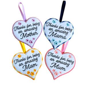Four felt heart hanging decorations embroidered with the message 'Thanks for being an amazing' followed by Mother, Mama, Mam, and Mum, each decorated with small flowers and ribbon loops for hanging.