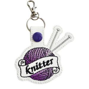 Faux leather keyring featuring an embroidered purple ball of yarn with two knitting needles and a banner reading 'knitter', finished with a purple snap fastener and attached to a metal keyring clip.