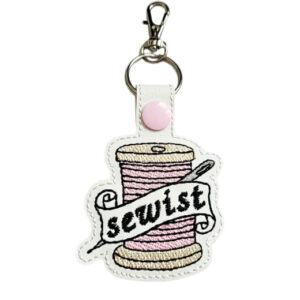 Faux leather keyring featuring an embroidered pink spool of thread with a needle and a banner reading 'sewist', finished with a pink snap fastener and attached to a metal keyring clip.