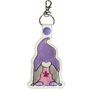 Little girl gnome keyring made from faux leather, featuring an embroidered gnome with a tall purple hat, long stitched hair, and a small flower on her dress, attached to a silver keyring ring and lobster clasp.