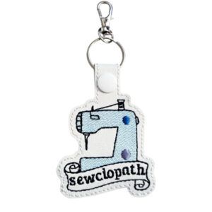 Faux leather keyring featuring an embroidered sewing machine with a banner reading 'sewciopath', finished with a white snap fastener and attached to a metal keyring clip.