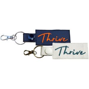 Two rectangular keyrings with the word "Thrive" embroidered on them in cursive lettering. One keyring is dark blue with orange embroidery, and the other is white with teal embroidery. Both keyrings have a metal clasp attached to a split ring.