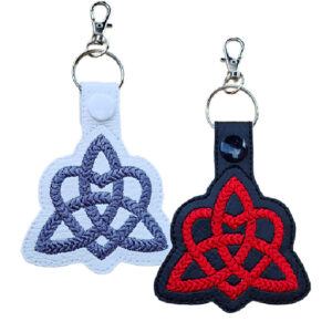 Two embroidered Trinity Heart Celtic Knot keyrings made from faux leather, one white with grey stitching and one black with red stitching, each with a snap button and metal clasp for attaching to keys or bags.