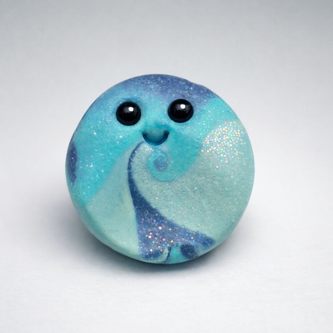 Handmade glow in the dark polymer clay Blob worry stone in swirling blue and turquoise with glitter, featuring a smiling face with round black eyes, calming pocket stone by Lollygag.