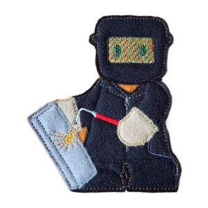 Felt gingerbread welder character wearing protective overalls, boots and a welding mask, holding a welding torch against a metal piece with stitched spark details.