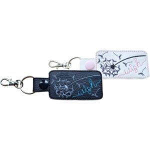 Two rectangular faux leather dandelion keyrings shown side by side. One is black with an embroidered dandelion and the word “wish” stitched in teal, and the other is white with an embroidered dandelion and the word “wish” stitched in light pink. Both keyrings have stitched edges, a strap with a snap button, and silver-toned lobster clasps with split rings.