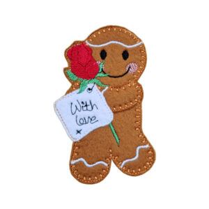 Handmade felt gingerbread character holding a red rose and a stitched tag reading “With love”, with embroidered icing details and a smiling face.