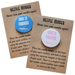 Two round needle minders displayed on kraft card packaging. One has a blue background with white text reading ‘Work In Progress’ and a thread illustration, and the other has a white background with pink text and the same thread design. Packaging reads ‘Needle Minder – Never lose your needle again!’ with usage instructions.