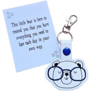 Bear face keyring made from white faux leather with embroidered facial details, brown ears and a pair of blue glasses, displayed on a printed keepsake card with the message ‘This little bear is here to remind you that you have everything you need to face each day in your own way.’