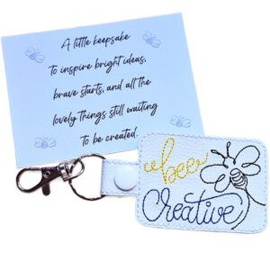 White faux leather Bee Creative keyring with embroidered bee design, shown with a matching keepsake card featuring an inspirational creative message.