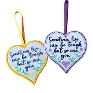 White felt heart hanging decorations with embroidered flowers and the message “Sometimes life can be tough but so are you”, shown with yellow and purple stitched borders and ribbon loops