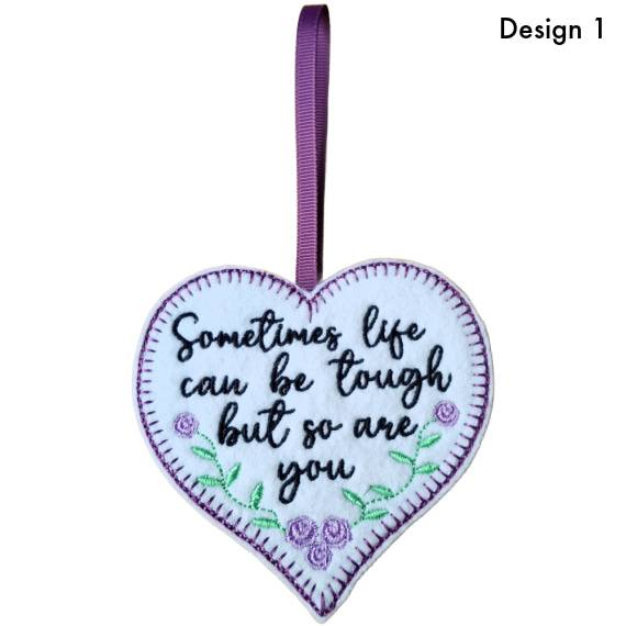 White felt heart hanging decoration with blanket stitch border, embroidered flowers and the message “Sometimes life can be tough but so are you”, finished with a purple ribbon loop (Design 1)