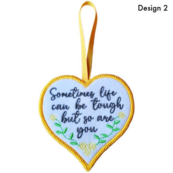 White felt heart hanging decoration with satin stitch border, embroidered flowers and the message “Sometimes life can be tough but so are you”, finished with a yellow ribbon loop (Design 2)