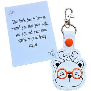Deer face keyring made from white faux leather with embroidered facial details, antlers and bright orange glasses, displayed beside a printed keepsake card with the message ‘This little deer is here to remind you that your light, your joy, and your own special way of being matter.’