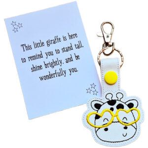 Giraffe face keyring made from white faux leather with embroidered facial details, black spots and bright yellow glasses, displayed beside a printed keepsake card with the message ‘This little giraffe is here to remind you to stand tall, shine brightly, and be wonderfully you.’