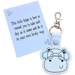 Hippo face keyring made from white faux leather with embroidered facial details and pale blue glasses, displayed beside a printed keepsake card with the message ‘This little hippo is here to remind you to take each day as it comes and do it in your own lovely way.’