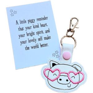 Pig face keyring made from white faux leather with embroidered facial details and pale pink heart-shaped glasses, displayed beside a printed keepsake card with the message ‘A little piggy reminder that your kind heart, your bright spirit, and your lovely self make the world better.’