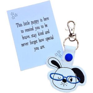 Puppy face keyring made from white faux leather with embroidered facial details, one black ear and bright blue glasses, displayed beside a printed keepsake card with the message ‘This little puppy is here to remind you to be brave, stay kind and never forget how special you are.’