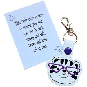 Tiger face keyring made from white faux leather with embroidered facial details, black stripes and bright purple glasses, displayed beside a printed keepsake card with the message ‘This little tiger is here to remind you that you can be both strong and soft, brave and kind, all at once.’
