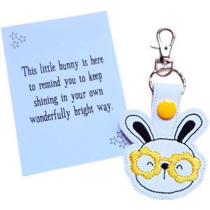 Smiley bunny keyring made from white faux leather with embroidered facial details, black ears and glittery gold glasses, displayed beside a printed keepsake card with the message ‘This little bunny is here to remind you to keep shining in your own wonderfully bright way.’