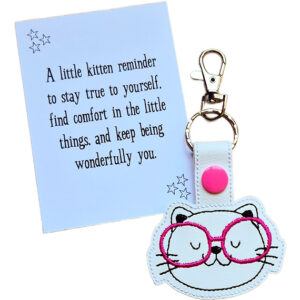 Kitten face keyring made from white faux leather with embroidered facial details, whiskers and bright pink glasses, displayed beside a printed keepsake card with the message ‘A little kitten reminder to stay true to yourself, find comfort in the little things, and keep being wonderfully you.’