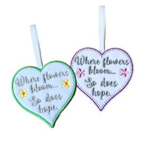 White felt heart hanging decorations with embroidered flowers and the message “Where flowers bloom… So does hope”, shown with green and purple stitched borders and ribbon loops