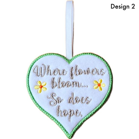 White felt heart hanging decoration with satin stitch border, embroidered flowers and the message “Where flowers bloom… So does hope”, finished with a white ribbon loop (Design 2)