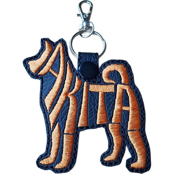 Black faux leather Akita word art keyring with orange embroidered lettering, black snap fastener and silver keyring clip.