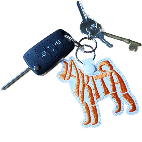White embroidered Akita word art keyring attached to a set of keys, showing its size and everyday use.