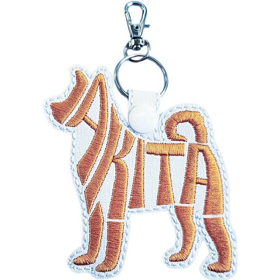 White faux leather Akita word art keyring with orange embroidered lettering, white snap fastener and silver keyring clip.