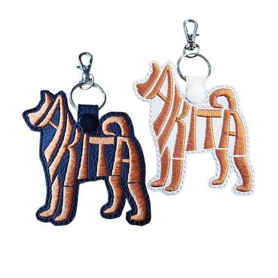 Two embroidered Akita word art keyrings in black and white faux leather, with silver clips and matching snap fasteners.