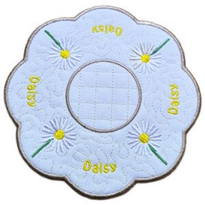 Close up of an embroidered April birth flower candle mat with white daisies, yellow centres, daisy text, stitched background detail and a scalloped border.