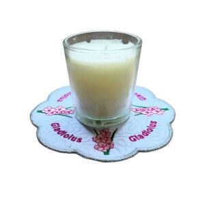 Embroidered August birth flower candle mat with pink gladiolus blooms, stitched text and a scalloped silver border, shown underneath a glass candle jar.