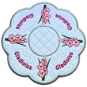 Close up of an embroidered August birth flower candle mat with pink gladiolus blooms, stitched gladiolus text, background detail and a scalloped silver border.
