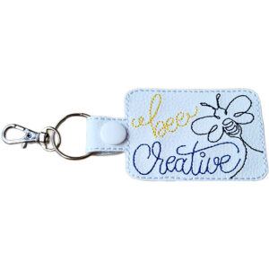 White faux leather Bee Creative keyring with embroidered bee design, yellow and black stitching, and silver metal clasp.