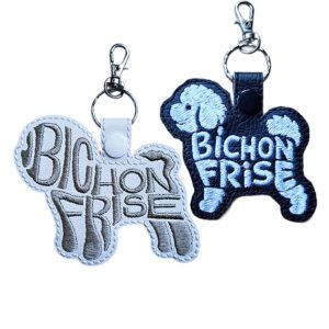 Two embroidered Bichon Frise word art keyrings in white and black faux leather, with silver clips and matching snap fasteners.