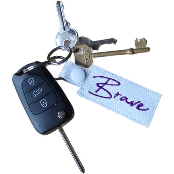 Embroidered Brave keyring attached to a set of keys, featuring white faux leather and the word 'Brave' stitched in purple.