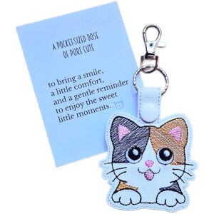 White faux leather calico cat keyring with embroidered kawaii cat design, shown with a matching keepsake card about pure cute and little moments.
