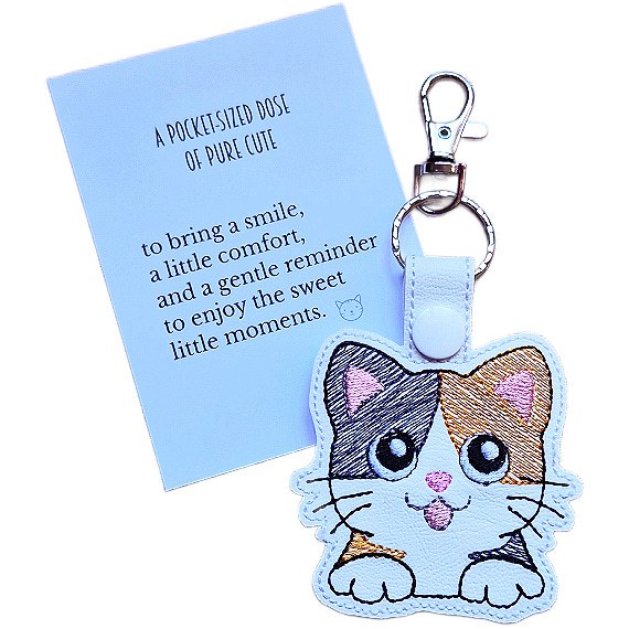 White faux leather calico cat keyring with embroidered kawaii cat design, shown with a matching keepsake card about pure cute and little moments.