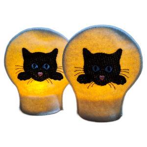 Lit embroidered cat LED tealight covers on white felt, glowing warmly with black cat faces and bright stitched details over battery operated tealights.