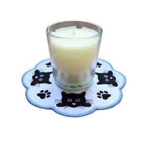 Embroidered cat paw print candle mat with black cat faces, stitched paw prints and a scalloped blue border, shown underneath a glass candle jar.