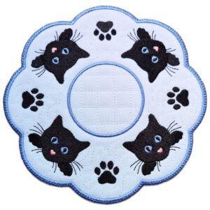 Close up of an embroidered cat paw print candle mat with black cat faces, stitched paw prints, a textured white background and a scalloped blue border.