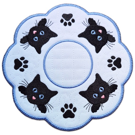 Close up of an embroidered cat paw print candle mat with black cat faces, stitched paw prints, a textured white background and a scalloped blue border.