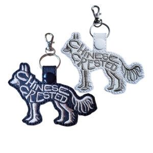 Two embroidered Chinese Crested word art keyrings in black and white faux leather, with silver clips and matching snap fasteners.