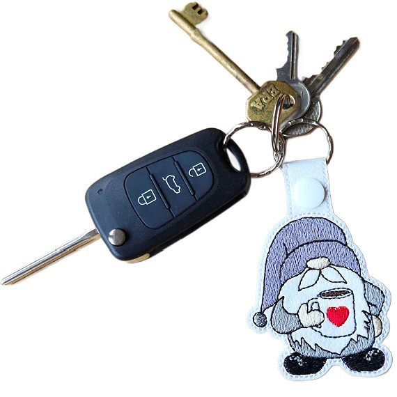 Embroidered coffee gnome keyring attached to a set of keys, featuring a cosy gnome holding a mug with a red heart.