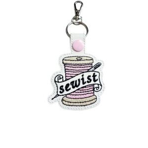 White faux leather Sewist keyring embroidered with a pink thread spool and needle design, finished with a metal clasp.