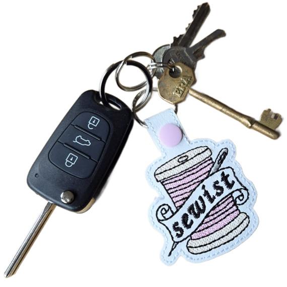 White faux leather Sewist keyring attached to a set of keys, embroidered with a pink thread spool and needle design.